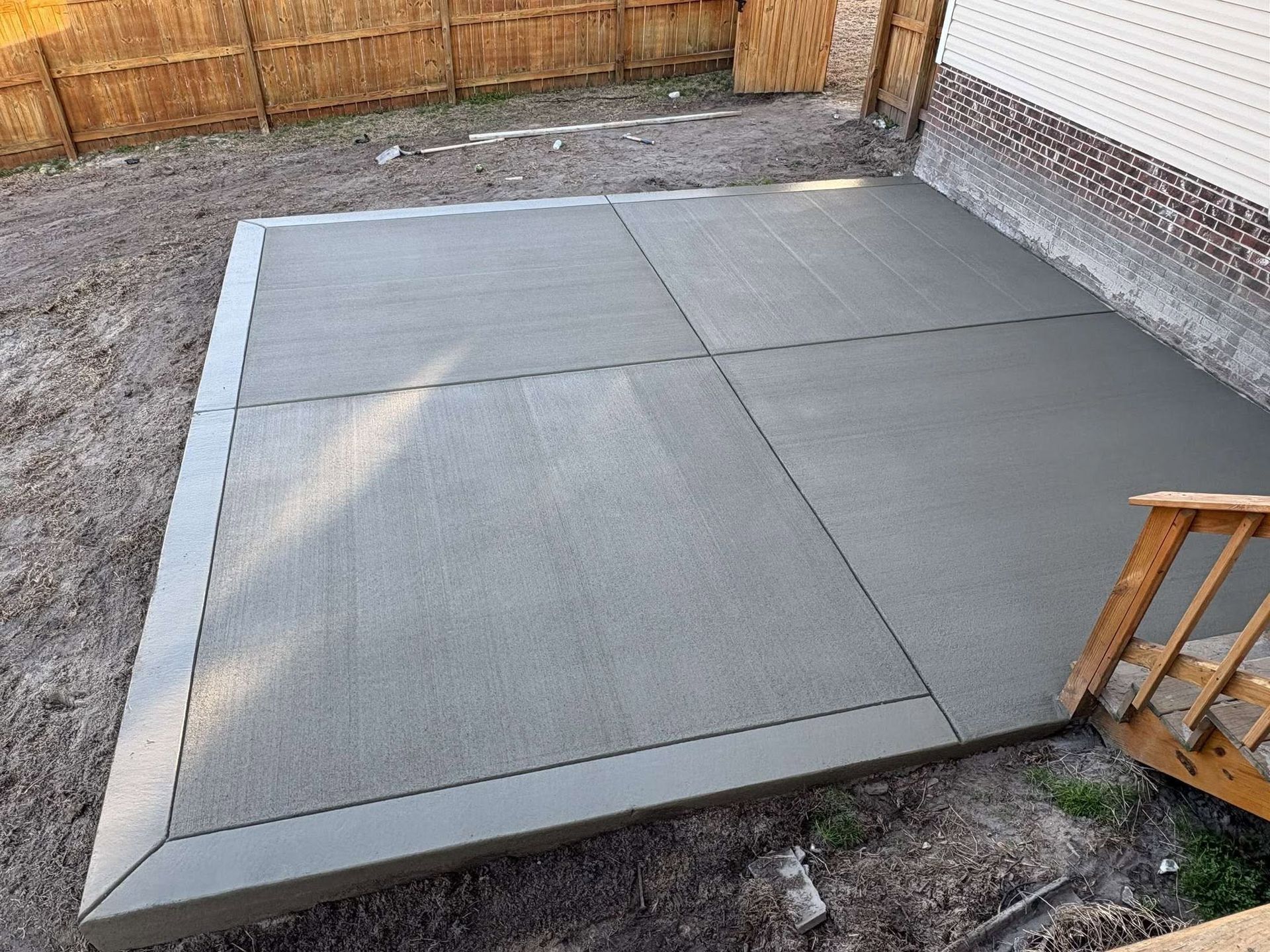 Freshly poured concrete patio slab in a fenced backyard, with wooden deck steps at the right.