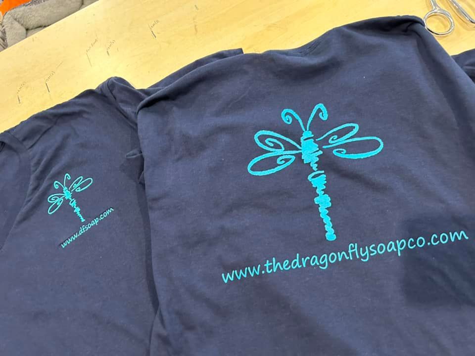 Two navy blue t-shirts displaying a cyan dragonfly logo and the text