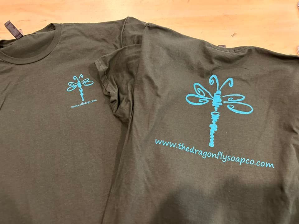 Two olive-green t-shirts, each featuring a light blue dragonfly logo and the website