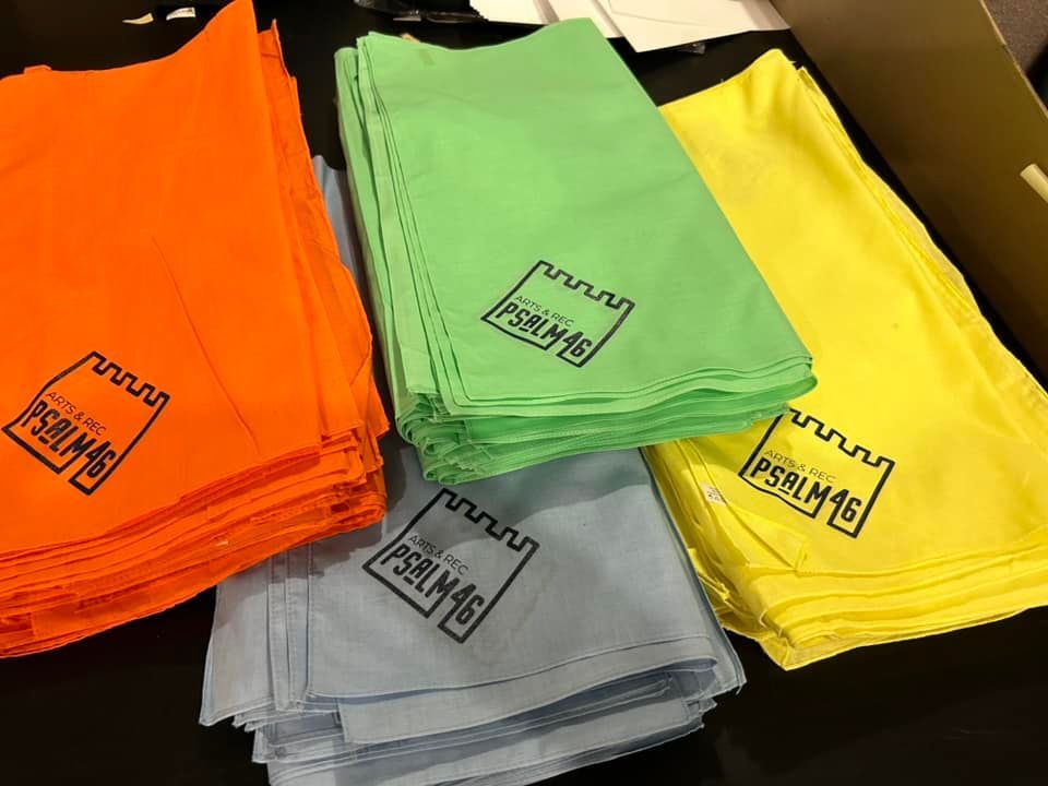 Four stacks of folded fabric squares in orange, green, yellow, and blue, each printed with a castle logo and PSALM 46.