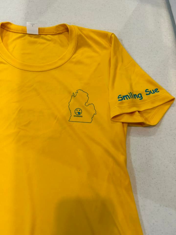A yellow t-shirt featuring a green outline of Michigan with a smiley face inside, and