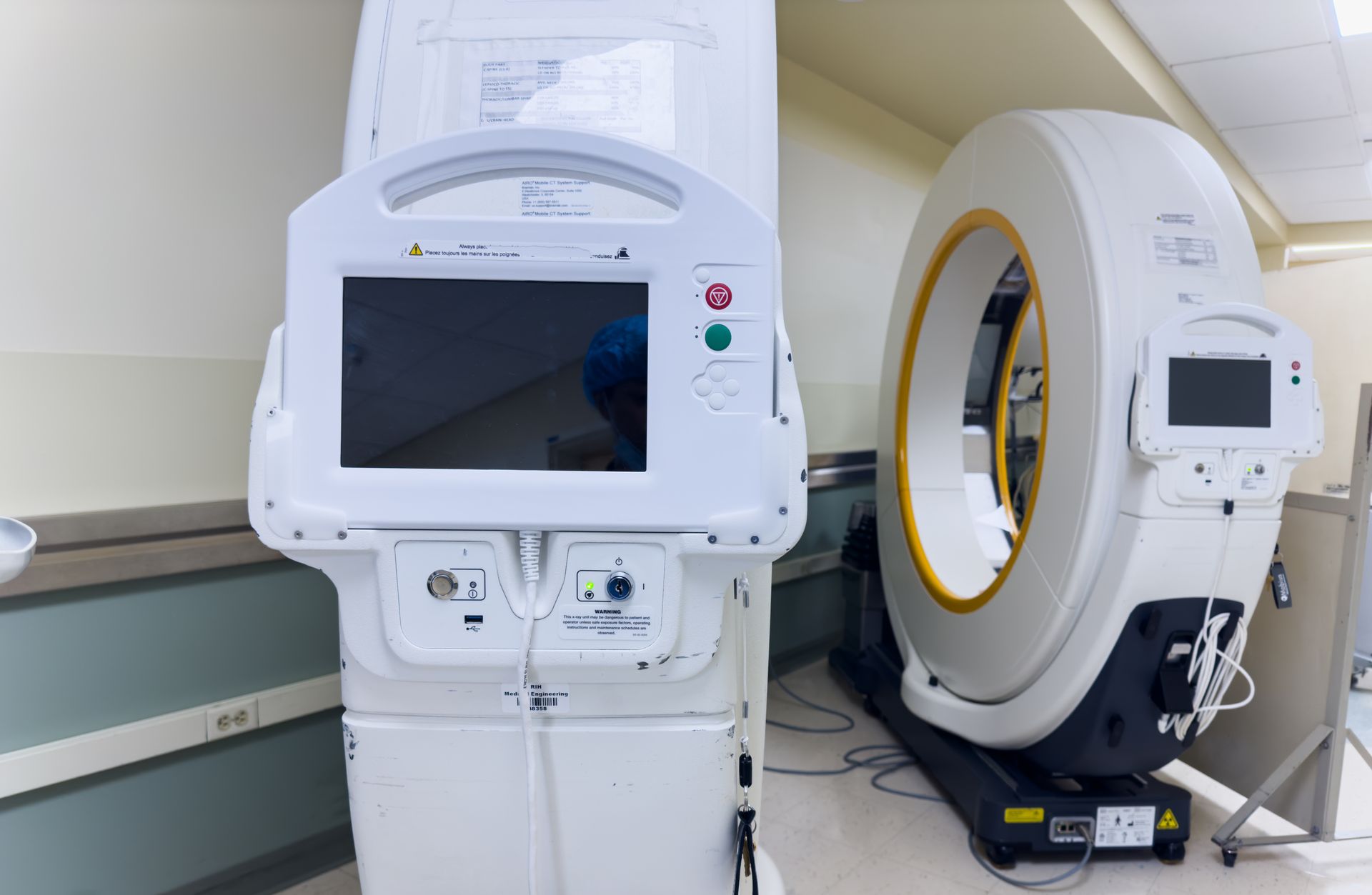 Medical imaging machines in a clinical room, including a standing monitor unit and a circular scanner.