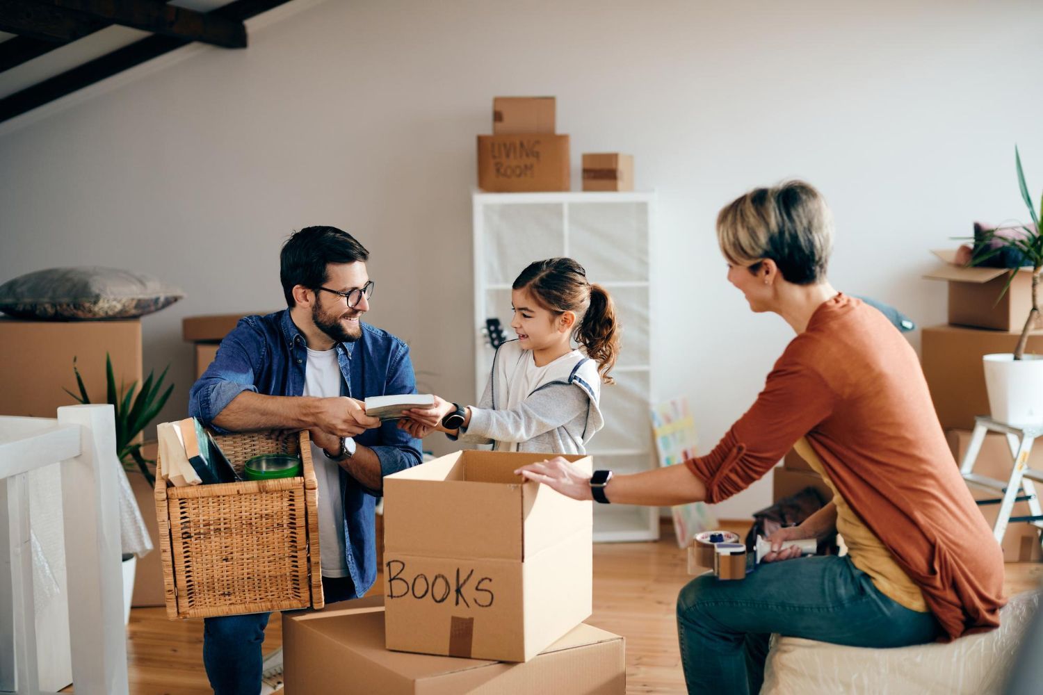 Family packing books into a box in a room with packed boxes; smiling father, daughter, and mother are working together.