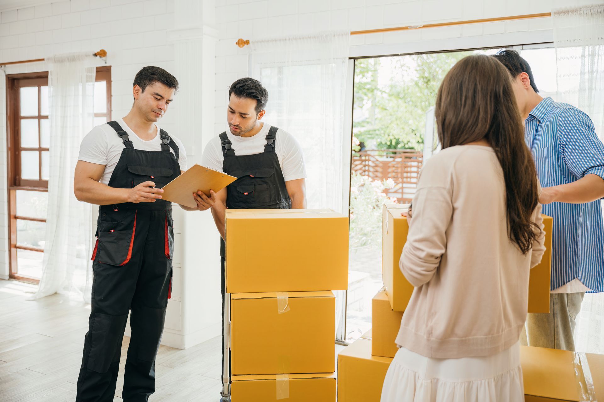 Two workers review paperwork beside stacked moving boxes in a bright room as a woman watches nearby