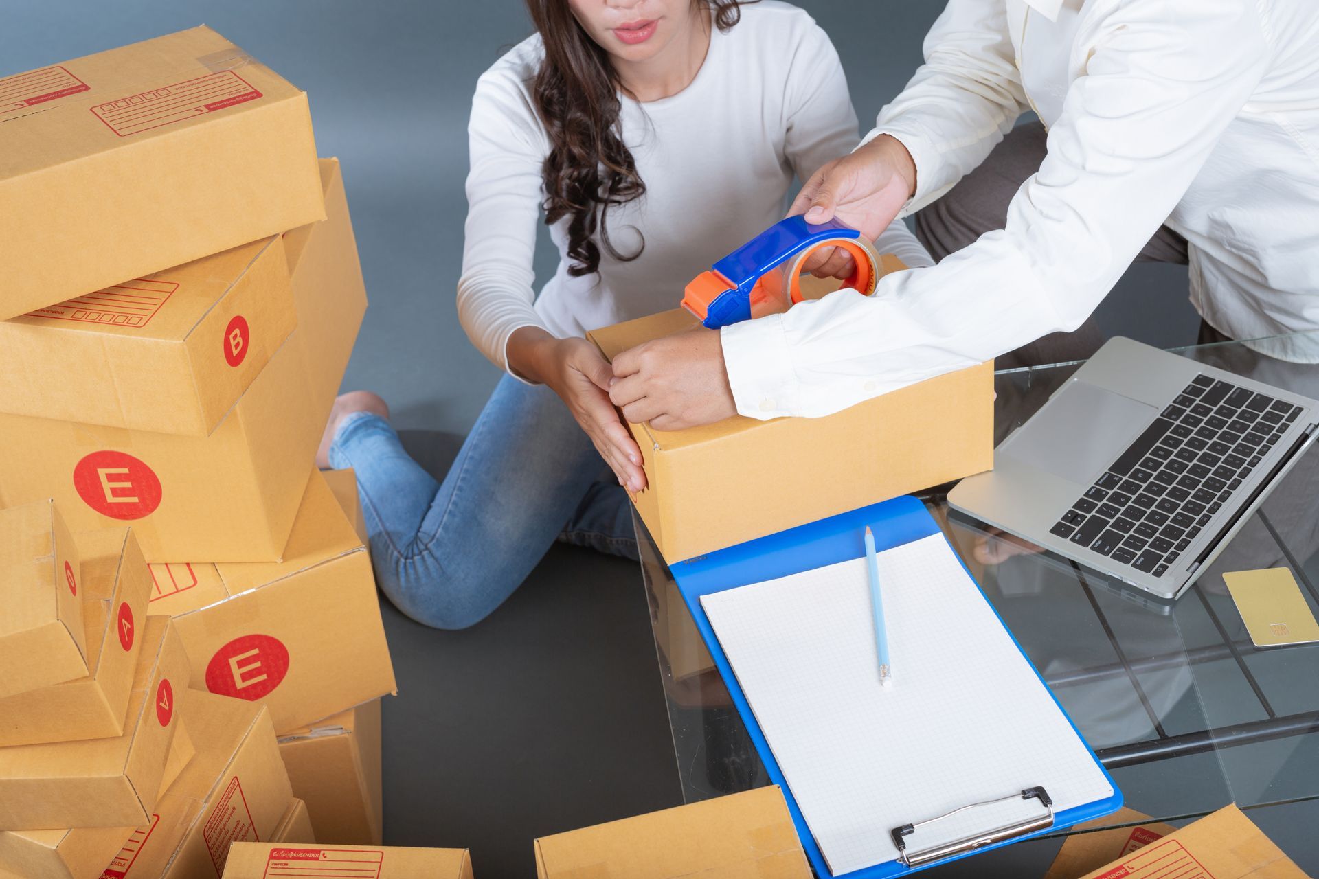 People packing boxes beside an open laptop and clipboard in a shipping workspace