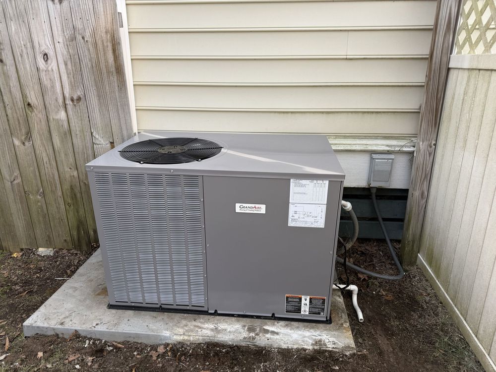 Outdoor central air conditioner unit beside a house wall and fence on a concrete pad.