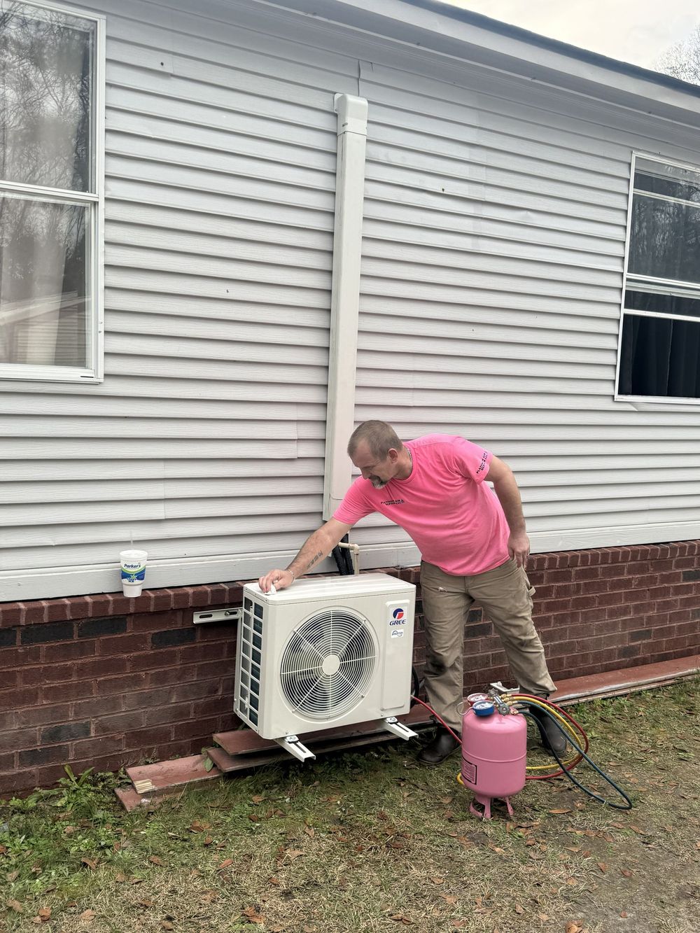 Person working on an outdoor air conditioner unit beside a house, with a pink propane tank nearby.