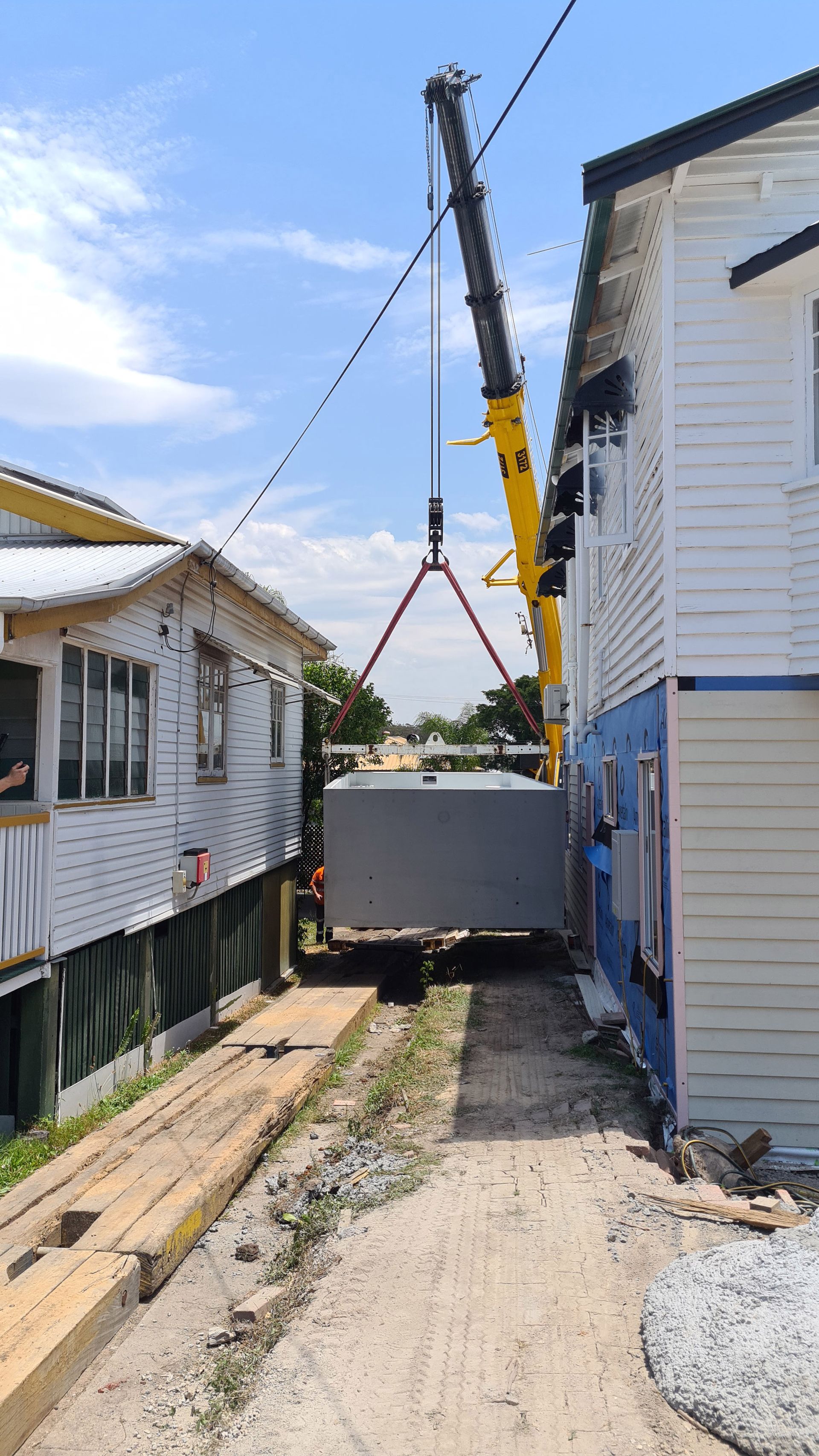 A crane lifts a large, grey box between two white houses. The houses are close together, with a narrow walkway in between.