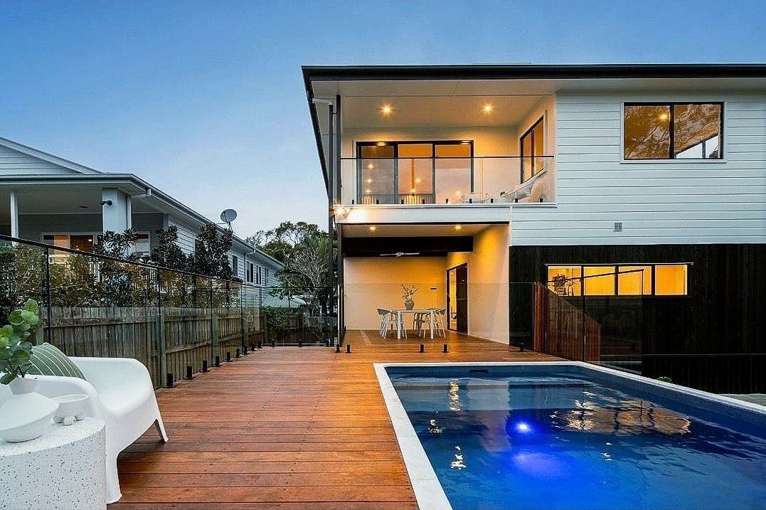 A two-story house with a pool and wooden deck. The pool water is blue, and there is outdoor seating.