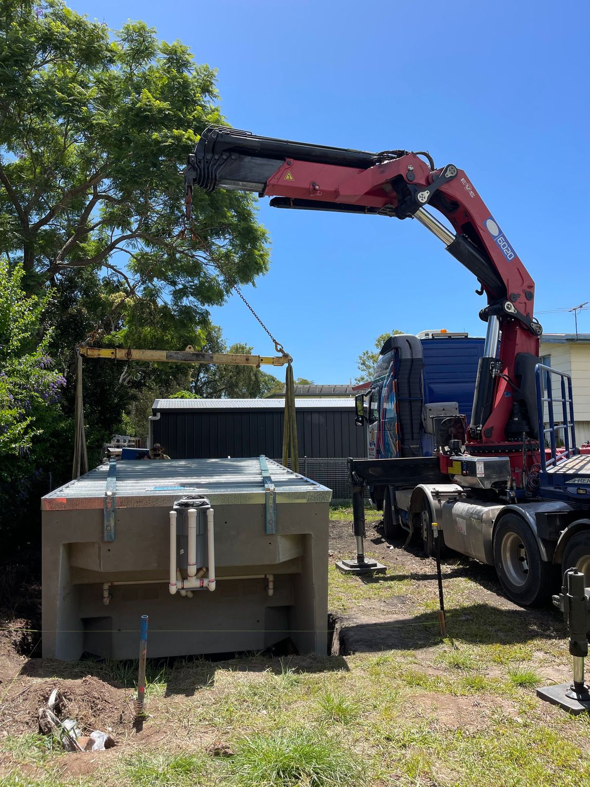 A large crane lifts a gray rectangular tank on a sunny day, likely for installation in a yard.