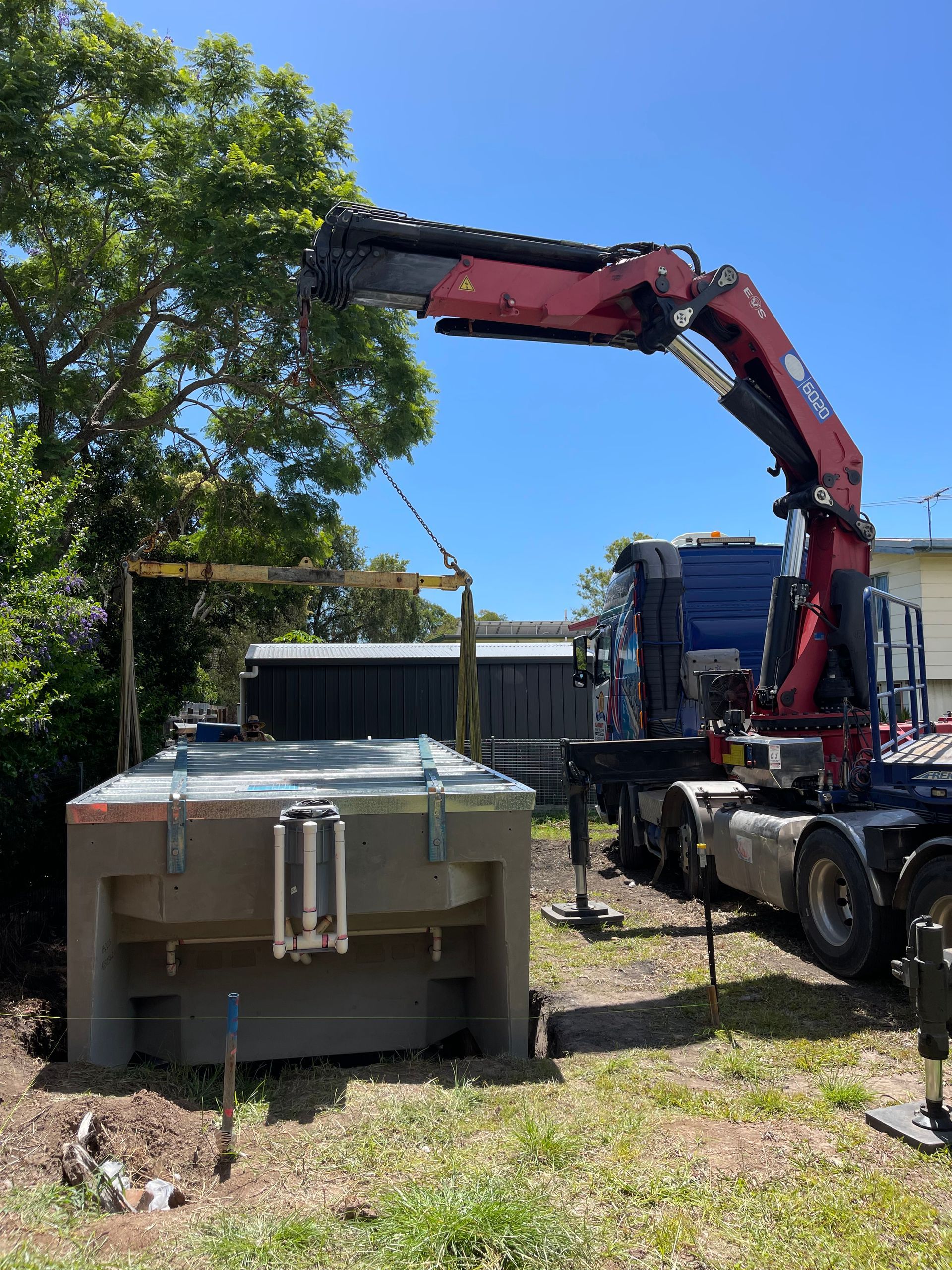 A large crane lifts a gray rectangular tank on a sunny day, likely for installation in a yard.