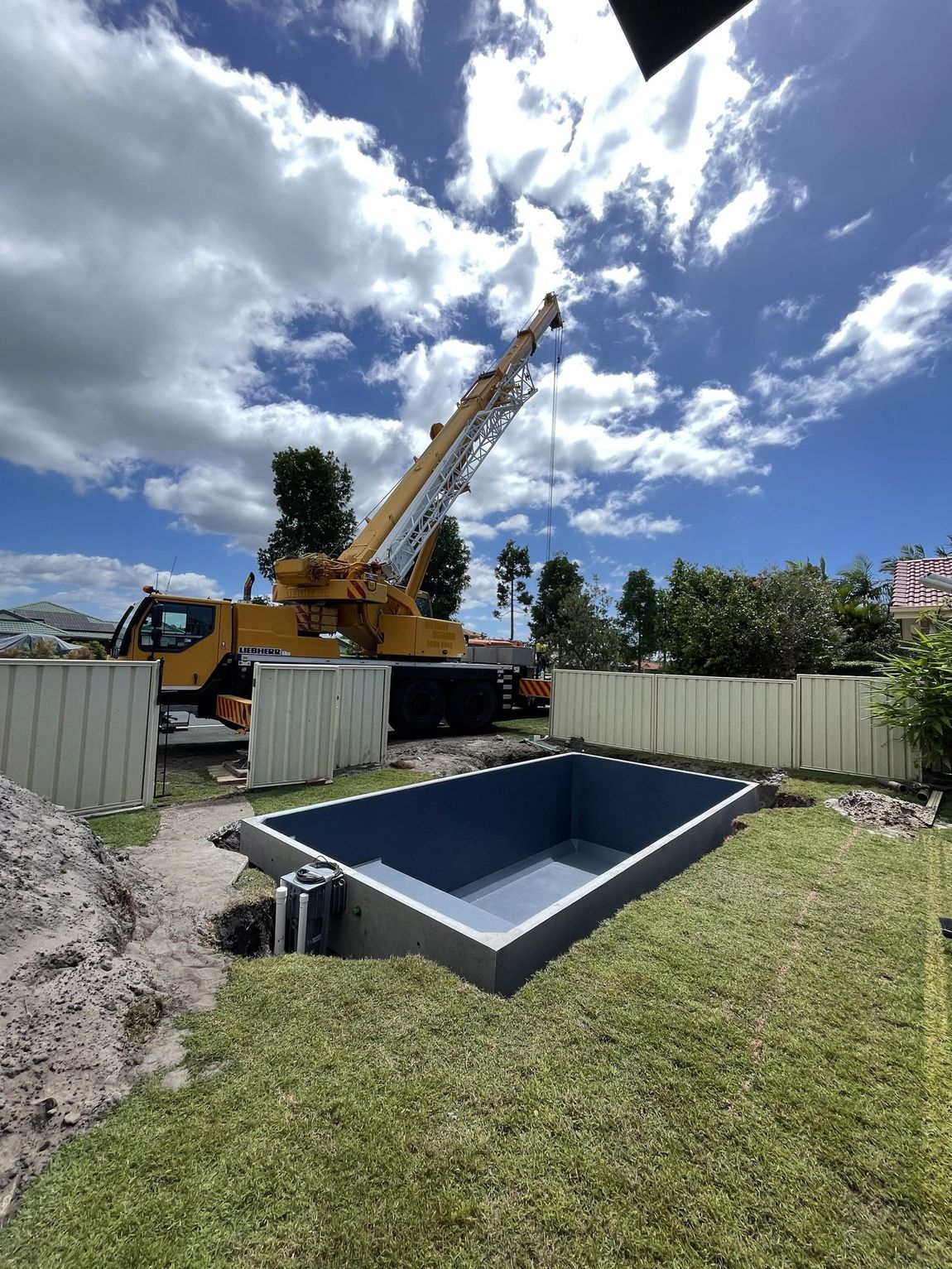 A crane lifting a rectangular, blue-lined swimming pool into a yard under a partly cloudy sky.