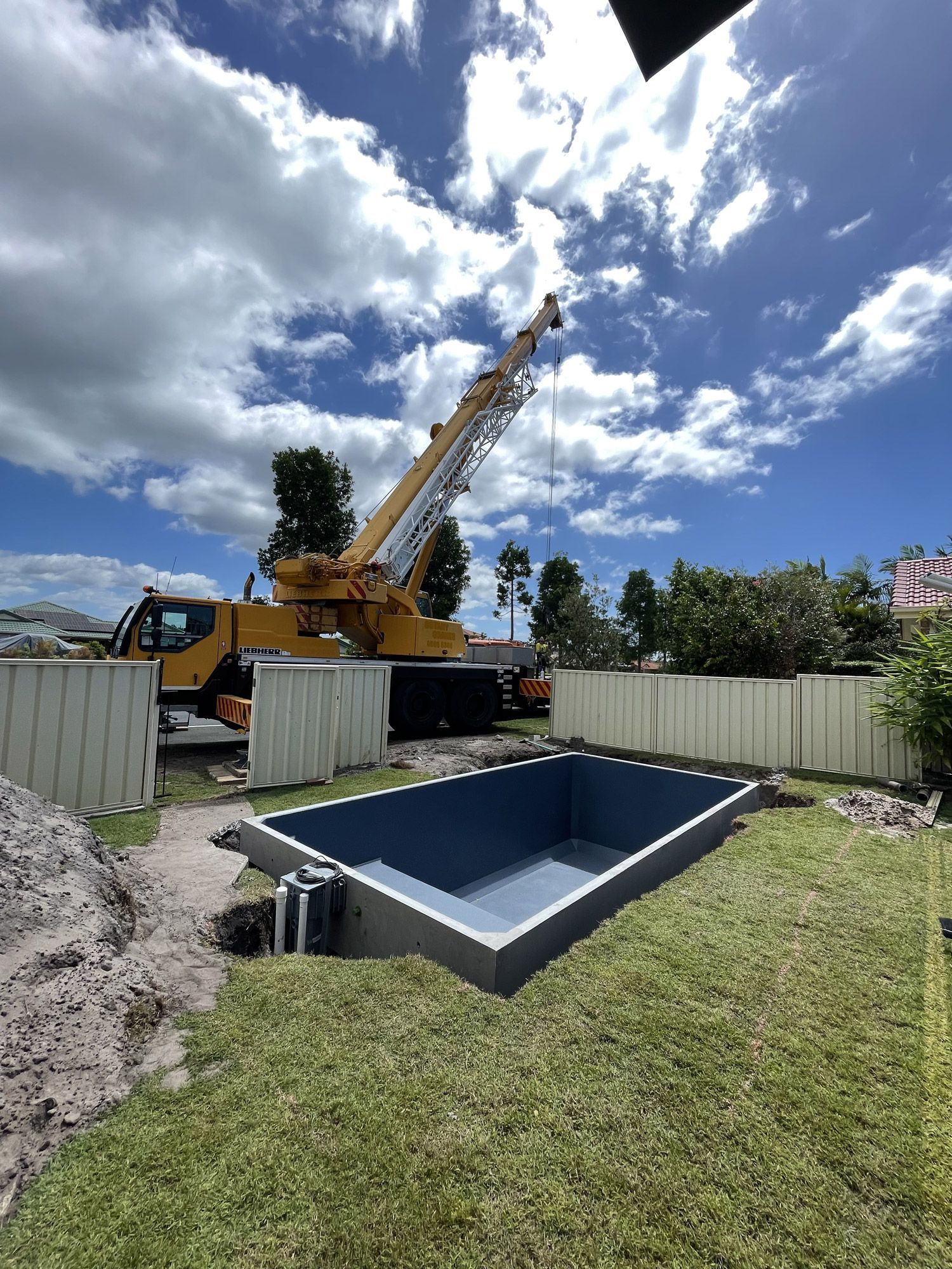 A crane lifting a rectangular, blue-lined swimming pool into a yard under a partly cloudy sky.