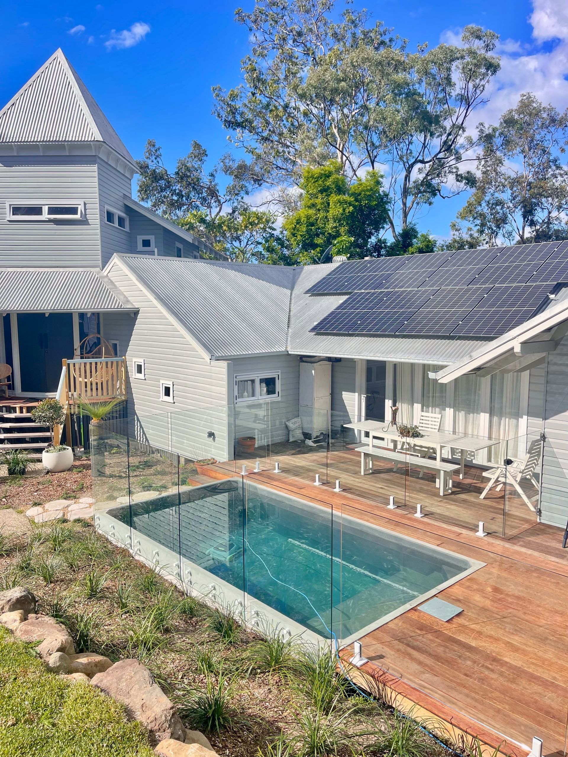 Exterior view of a light grey house with a pool, solar panels, and wooden deck. Trees and blue sky in the background.