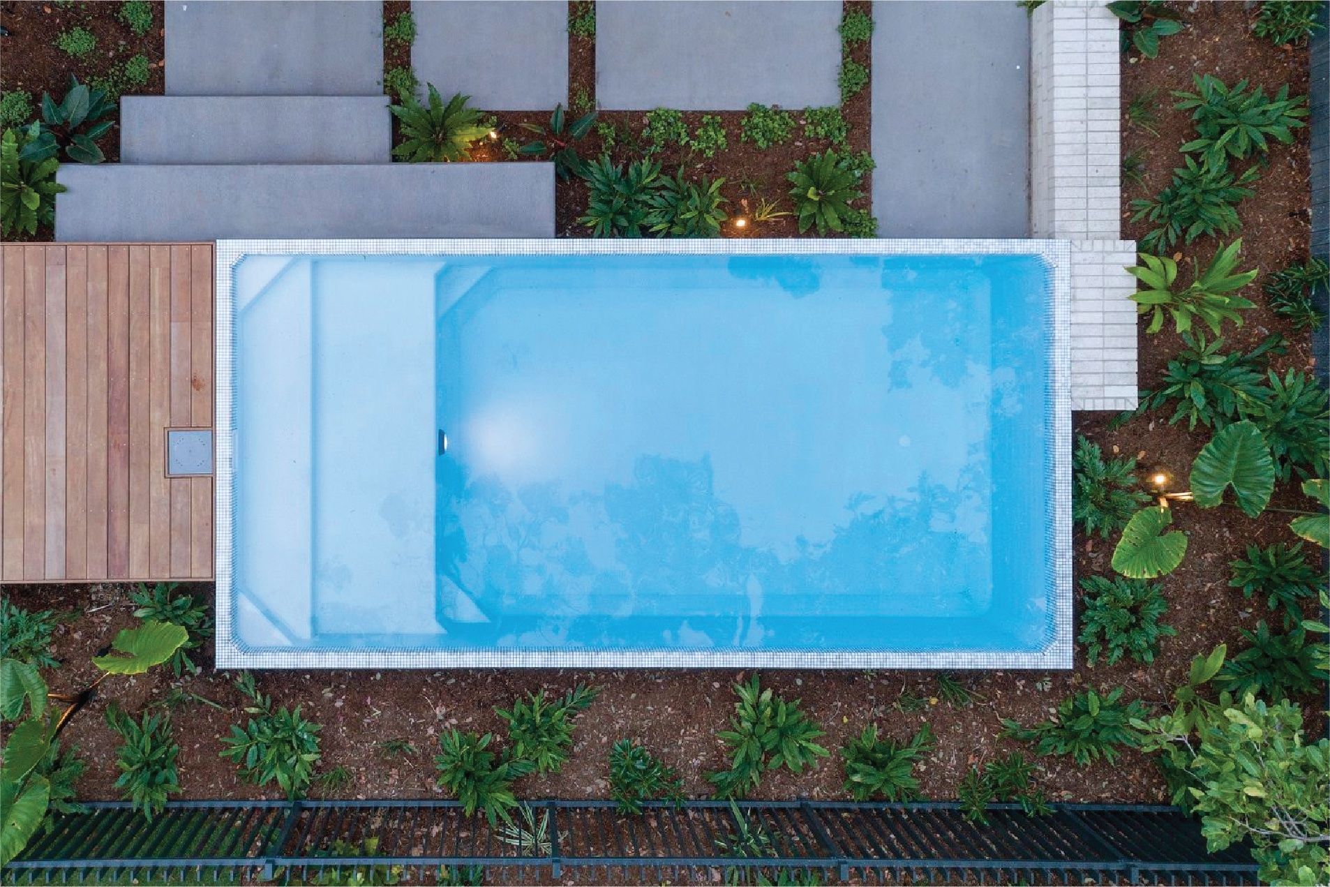 Overhead view of a rectangular pool surrounded by a wooden deck, concrete steps, and lush greenery in a residential backyard.