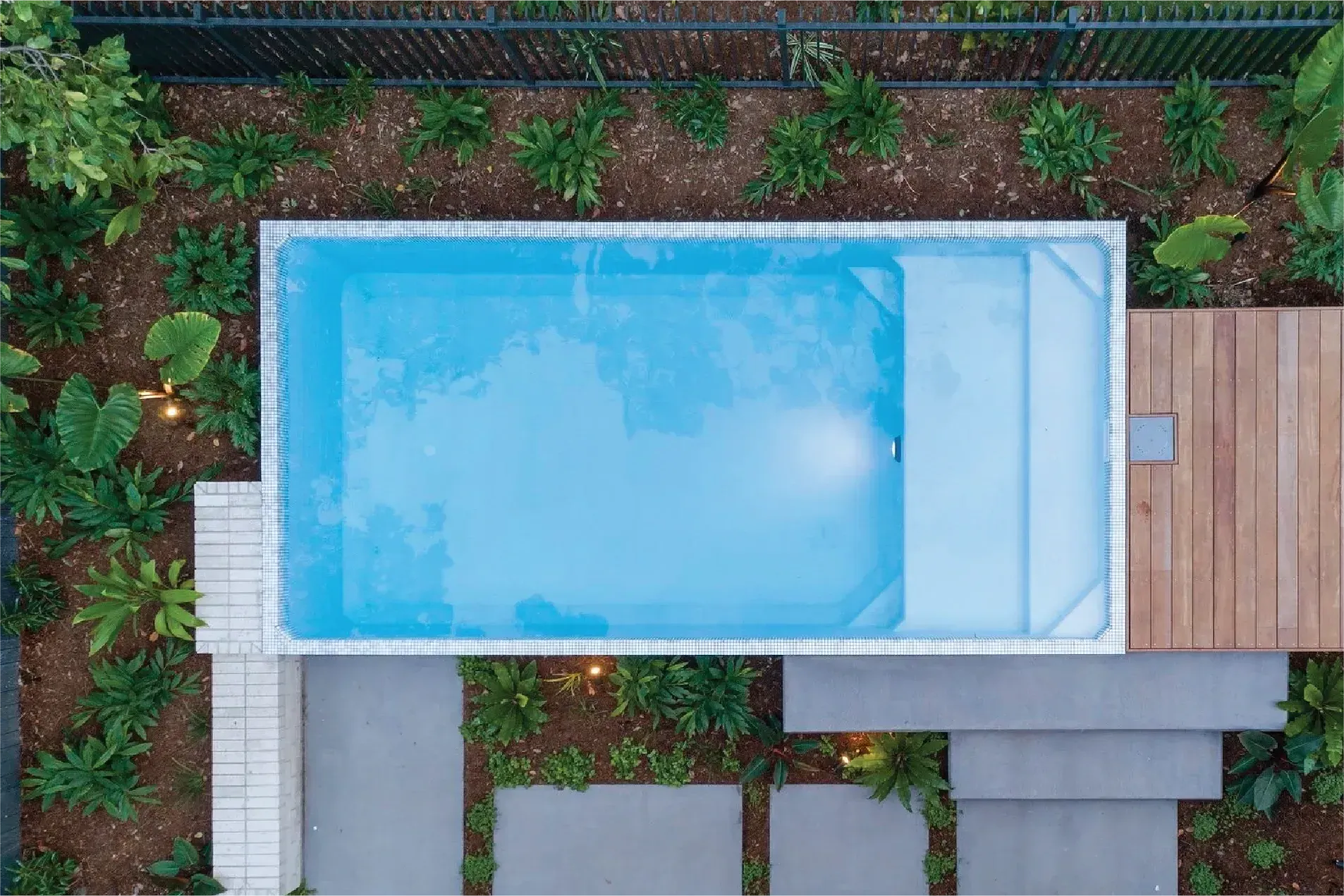 Overhead view of a rectangular pool surrounded by greenery, with a wooden deck and stone pathway leading to it.