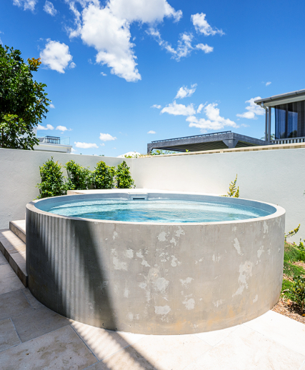 Circular concrete plunge pool with blue water, against a white wall under a blue sky with clouds.