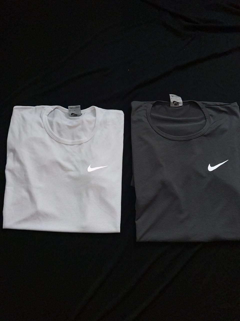 Dos camisetas deportivas Nike dobladas, una blanca y otra gris oscuro, sobre fondo negro.