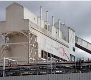 A large, white industrial building with several tall vents, metal support structures, and a logo on the side.