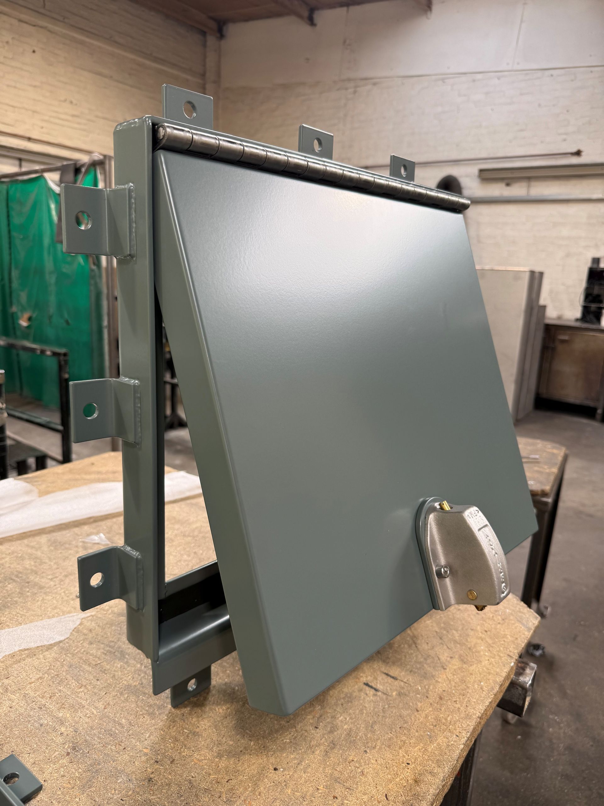 A gray metal access panel door with a locking latch, shown in an open position on a workshop table.