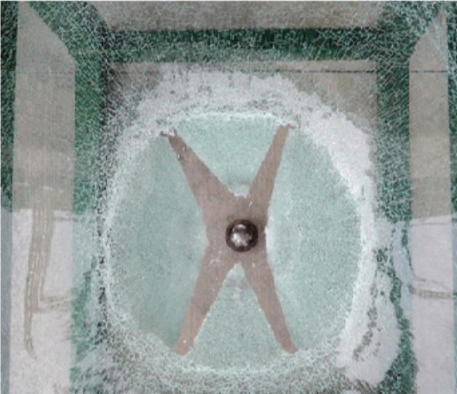 An overhead view of cracked, tempered glass pane with an X-shaped metal support bracket centered around a bolt.