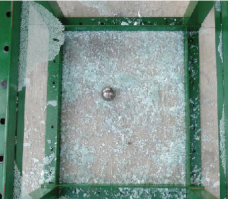 Shattered tempered glass fragments contained within a green metal frame, with a single metal ball resting in the center.