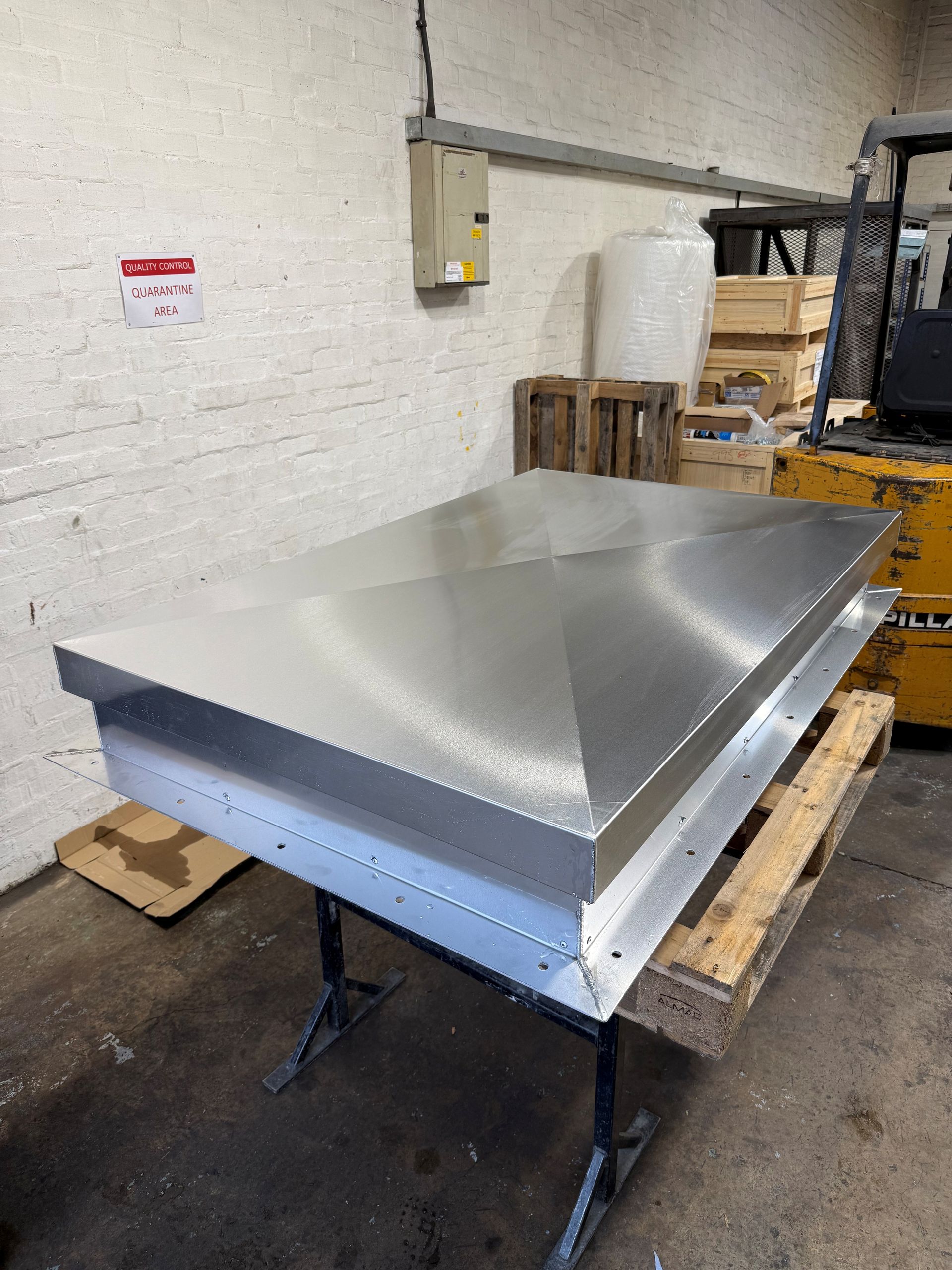 A metallic, rectangular roof vent cover with a peaked top sits on a workbench inside a warehouse.