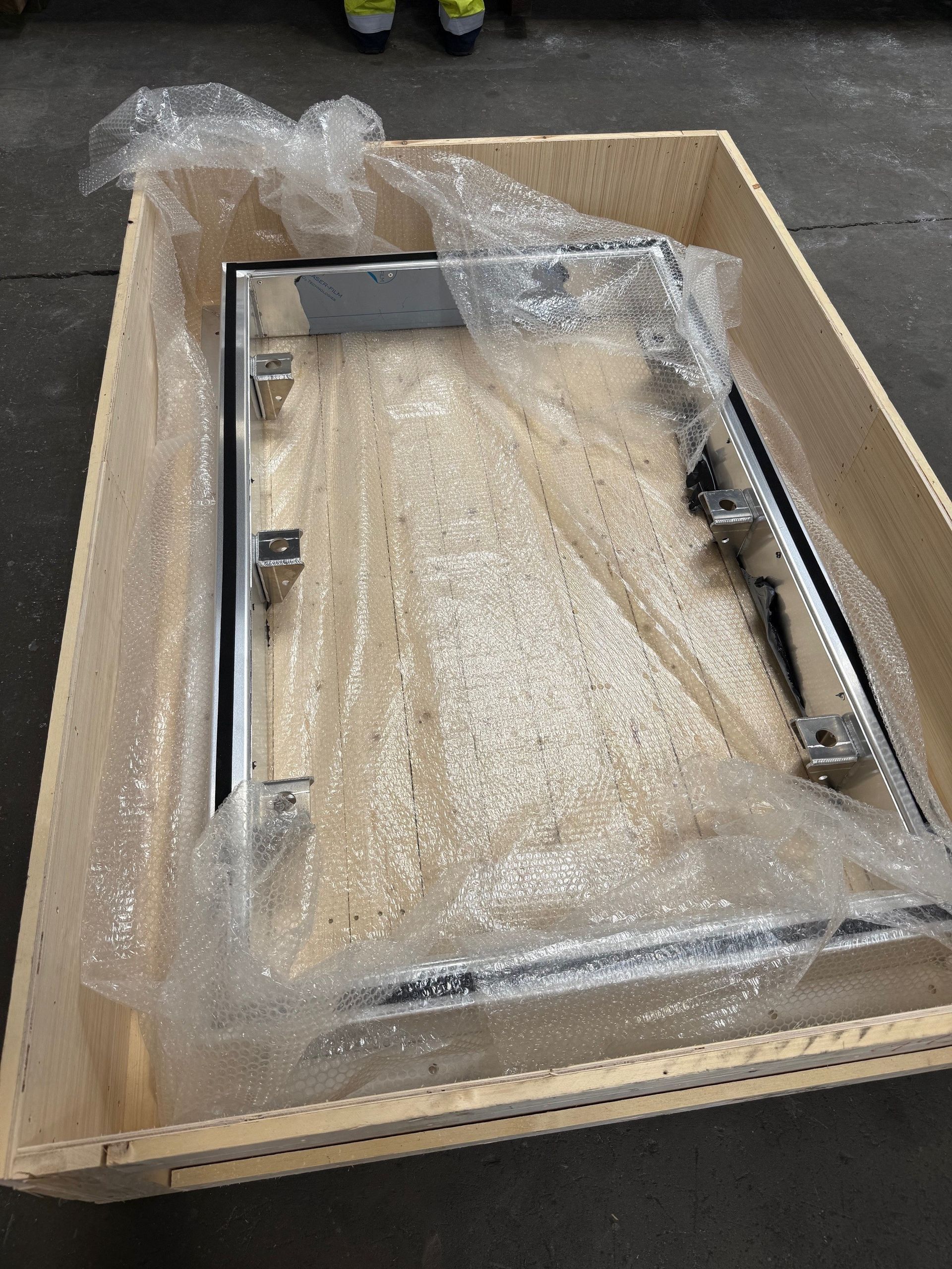 A rectangular metal frame sits inside a wooden crate, partially covered by bubble wrap.