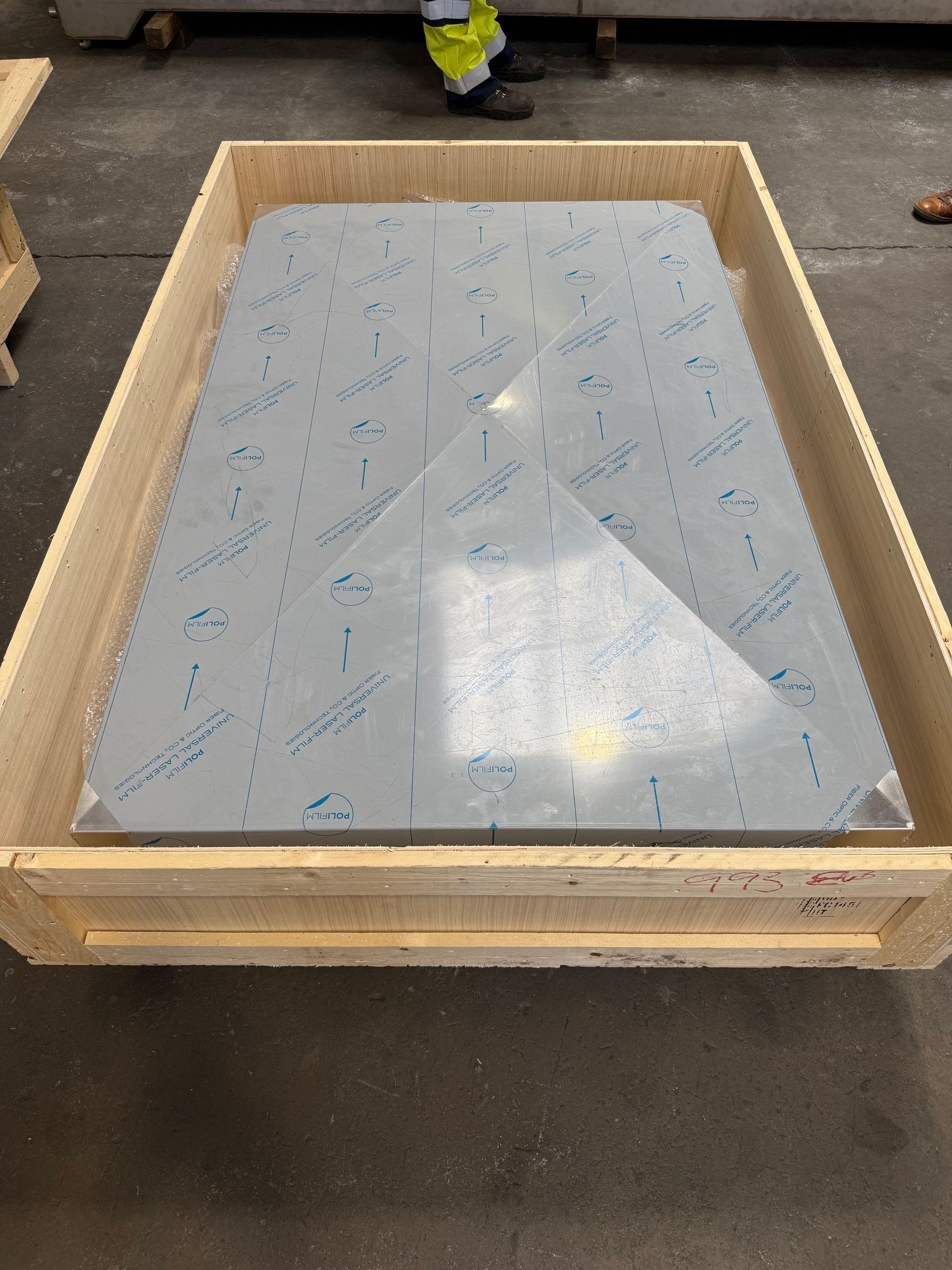 A large rectangular metal sheet covered in protective film sits inside a wooden shipping crate on a factory floor.