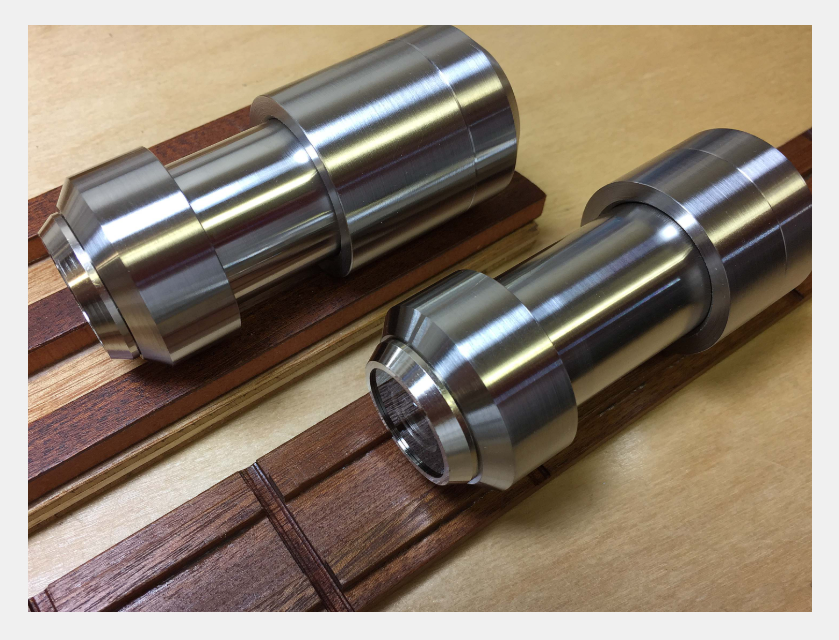 Two cylindrical machined metal components resting on dark wooden parallel strips on a light-colored surface.