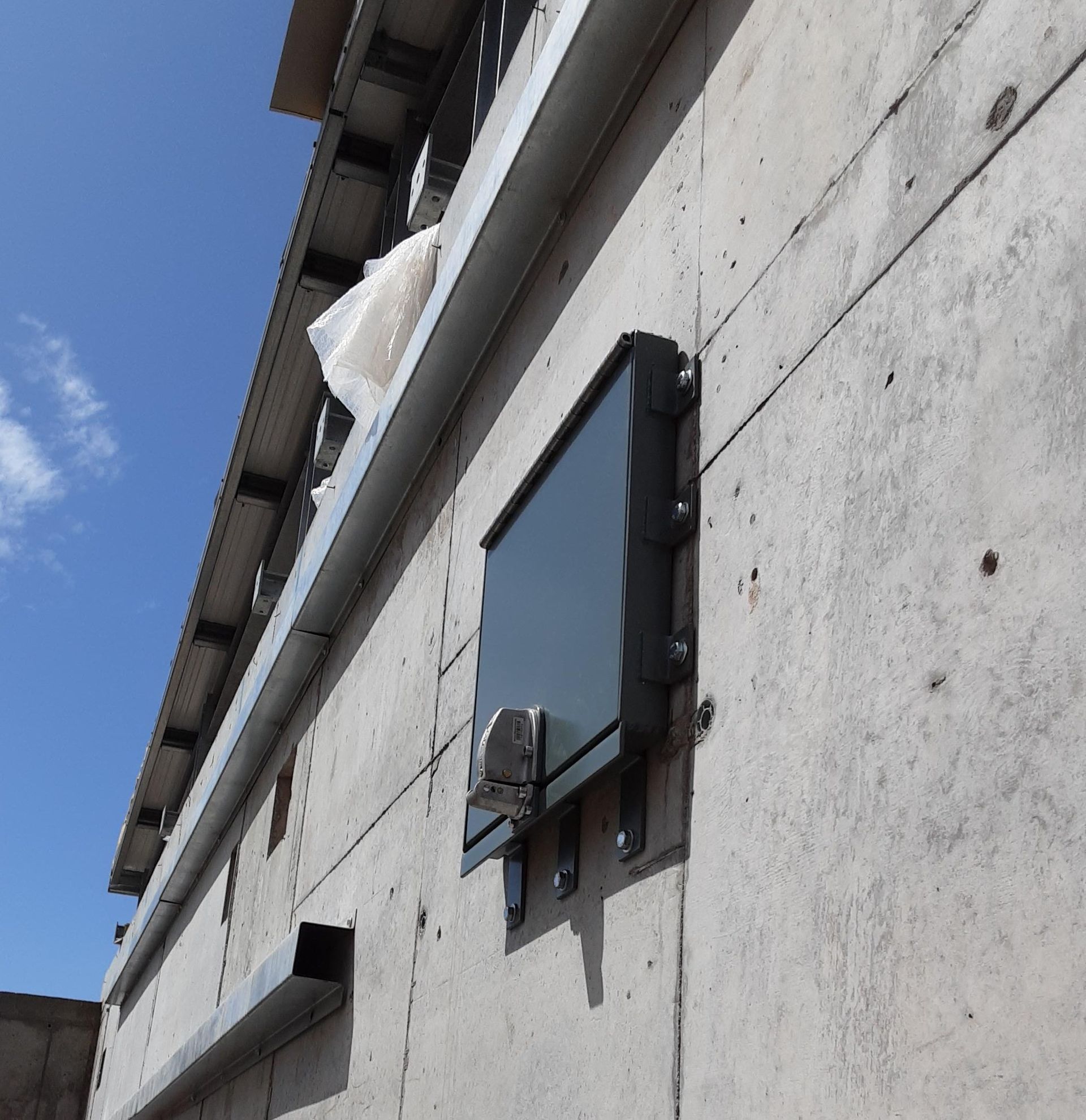A dark, rectangular utility box is mounted to a textured concrete exterior wall beneath a roof overhang.