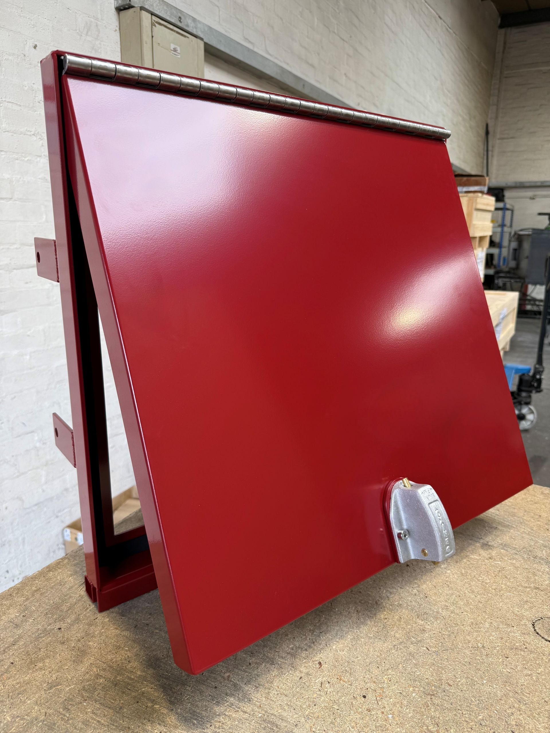 A red, hinged metal access door or hatch with a silver latch, shown from a slightly elevated angle in an industrial space.