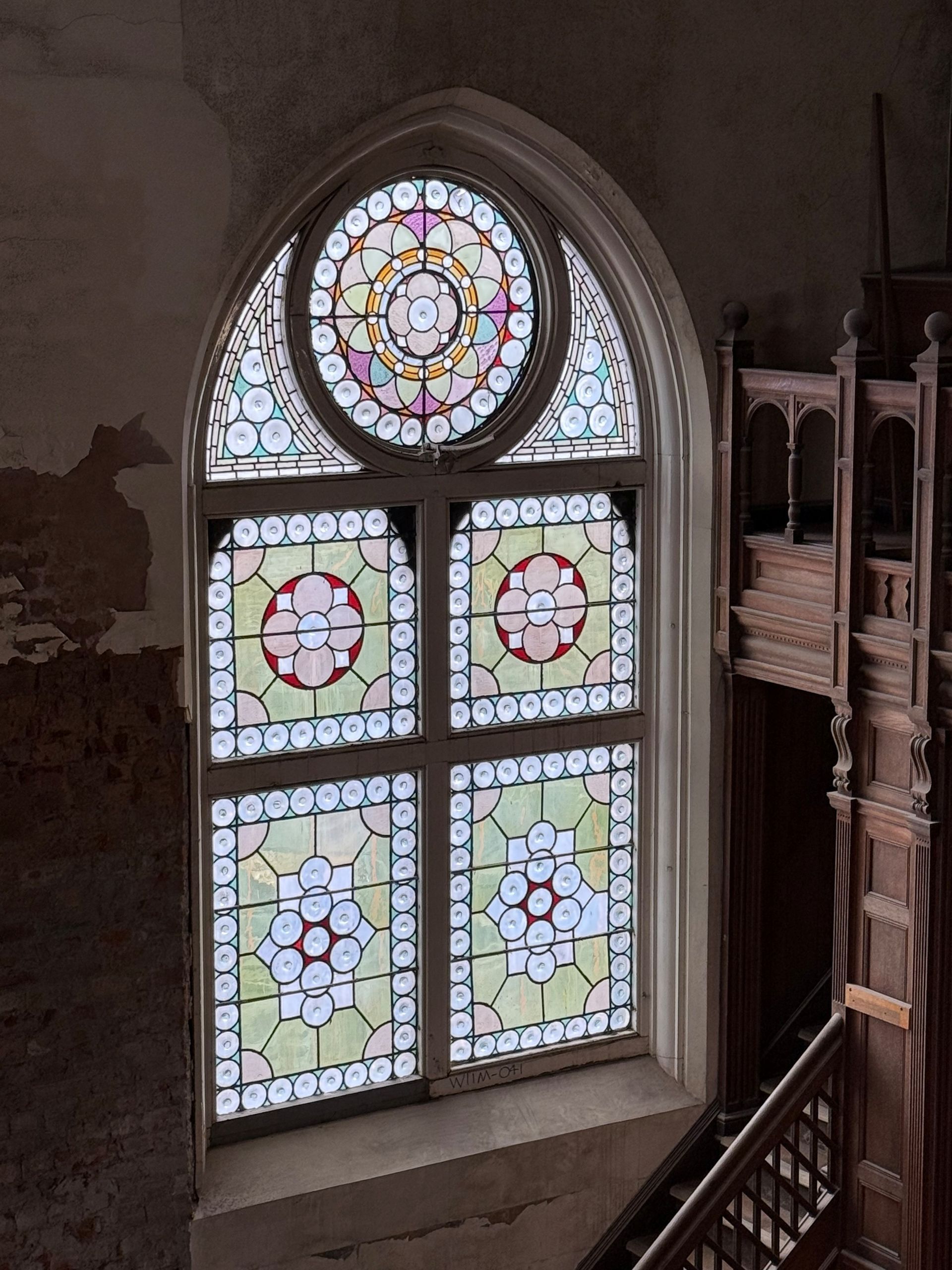 A stained glass window with a circular top section, set in a stone wall next to a dark wood balcony and staircase.