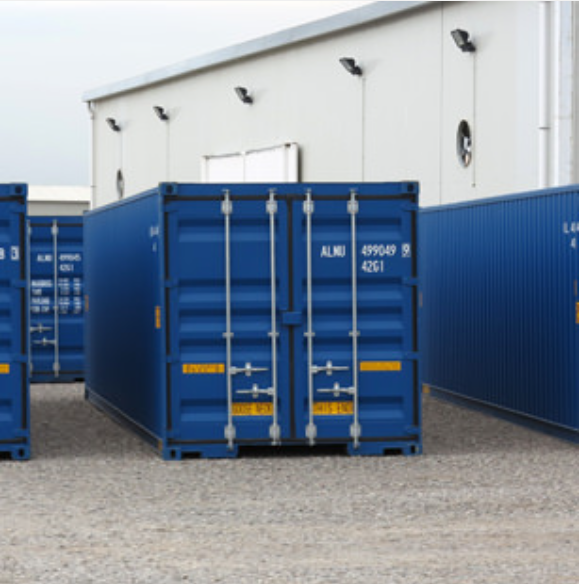 Three blue shipping containers sit on a gravel ground in front of a white warehouse building.