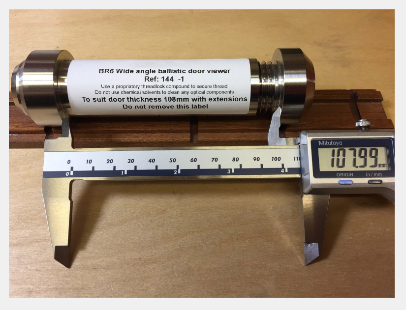 A wide-angle door viewer, measuring 107.99 mm in length, is being measured by digital calipers on a wooden surface.