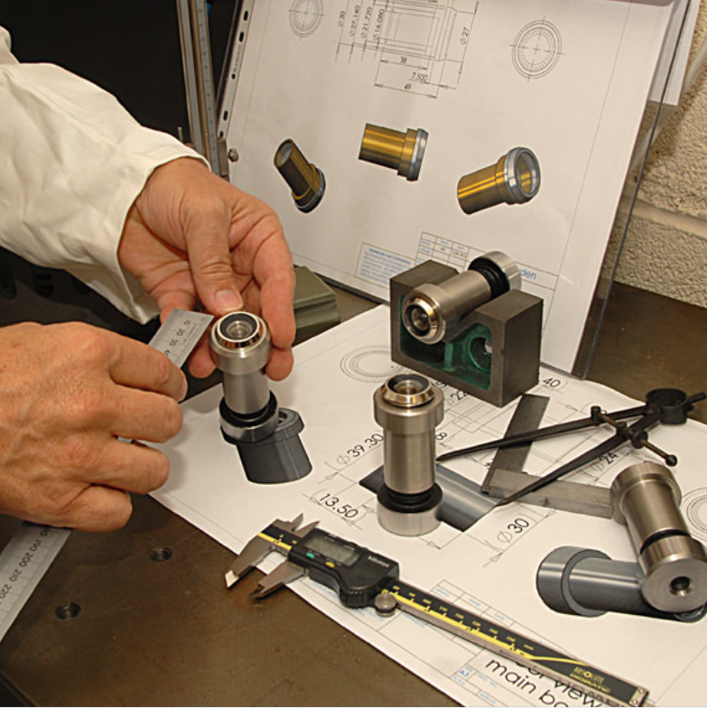 A person inspecting metal mechanical parts using a ruler, digital calipers, and technical blueprints on a workshop table.