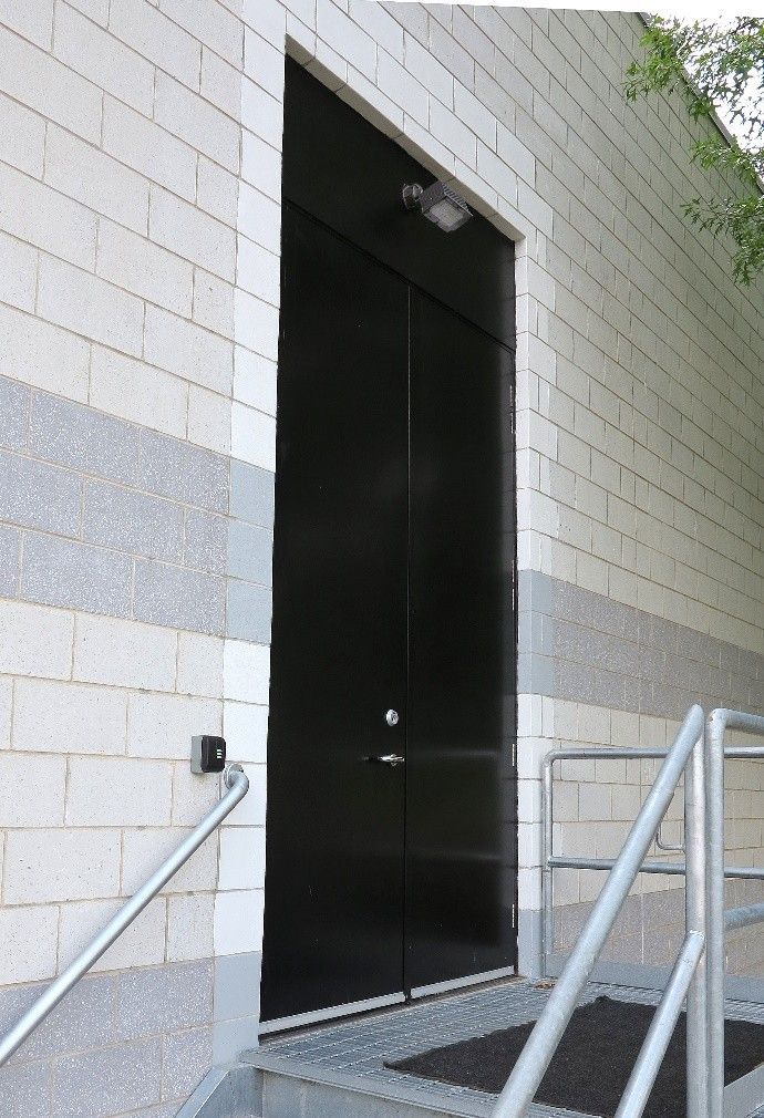 A tall black double door set in a light-colored brick wall with a metal handrail and stairs leading to the entrance.