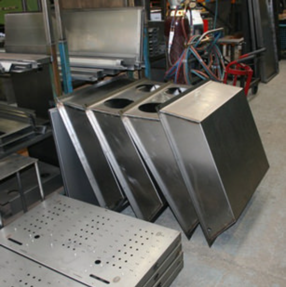 Stacks of metal fabrication components in a workshop, including sheet panels with perforations and rectangular casings.