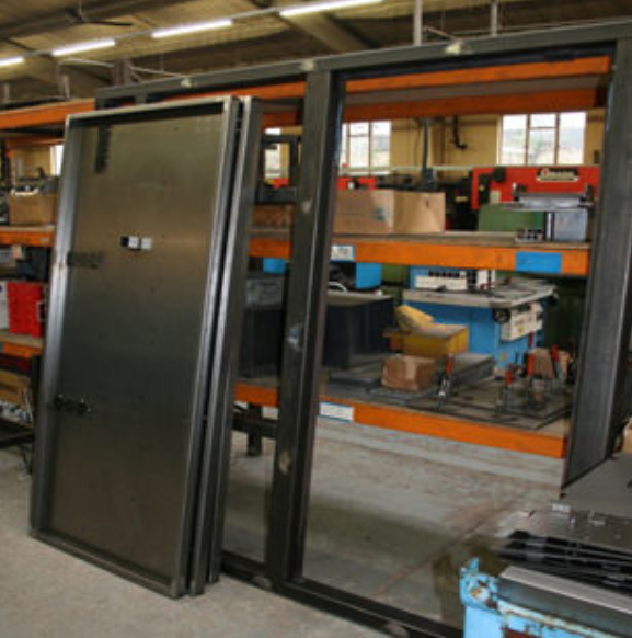 A metal door frame and panel standing in a workshop with industrial shelving and machinery in the background.