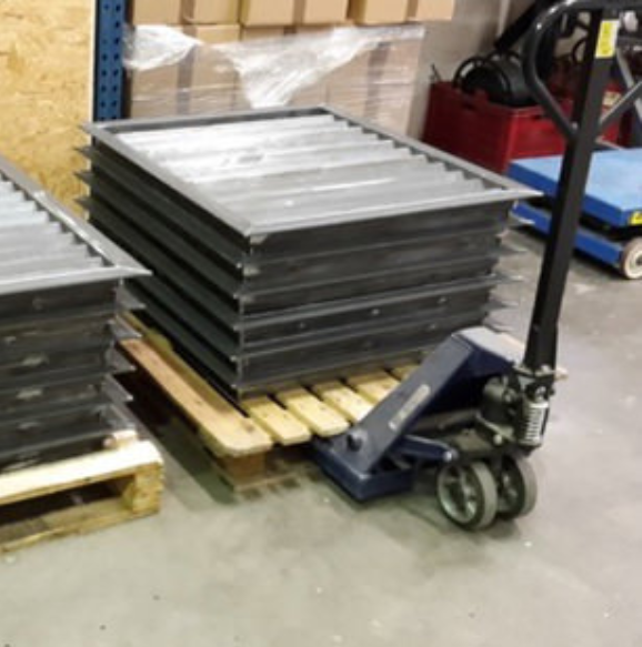 A pallet jack moves a stack of rectangular metal ventilation grilles on a wooden pallet in a warehouse.