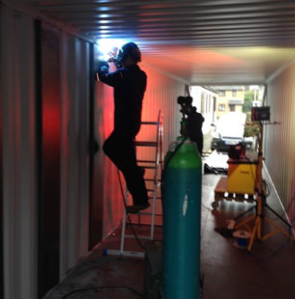 A person in protective gear welds a metal panel inside a container, standing on a ladder near a green gas cylinder.