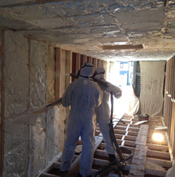 Two workers in protective suits spray insulating foam onto the walls and ceiling of a wooden frame structure.