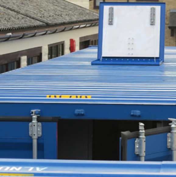 A blue shipping container roof with a raised ventilation hatch, positioned in front of a building with a tiled roof.
