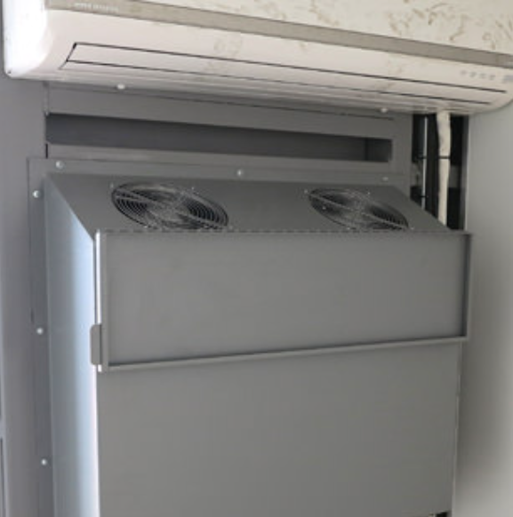 A grey metal server cabinet enclosure mounted on a wall with two circular exhaust fans, located below an AC unit.
