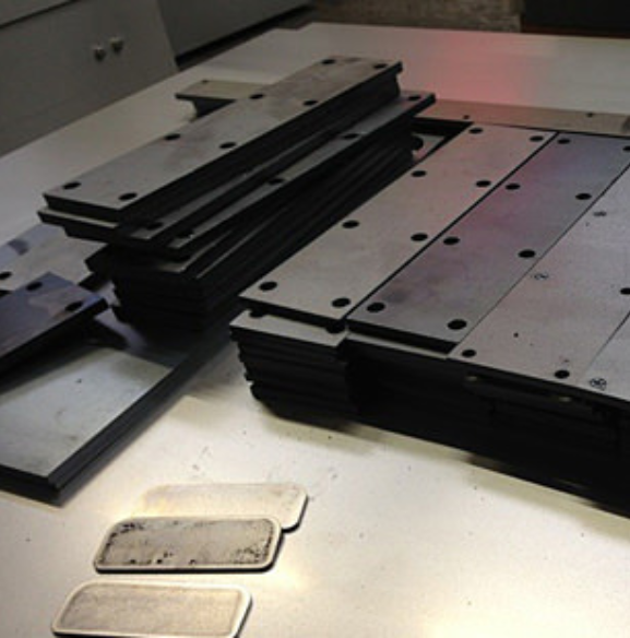 Stacks of dark, rectangular metal plates with pre-drilled holes arranged on a workbench, with smaller metallic parts below.