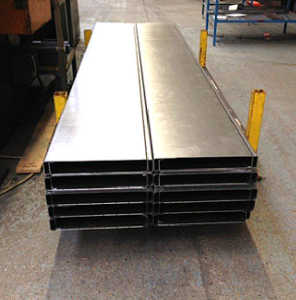 A stack of long, flat, rectangular metal industrial panels secured by yellow supports in a workshop setting.