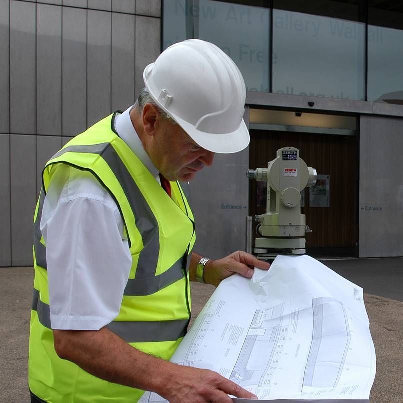 A person wearing a white hard hat and yellow high-visibility vest reviews blueprints next to a surveyor's level.