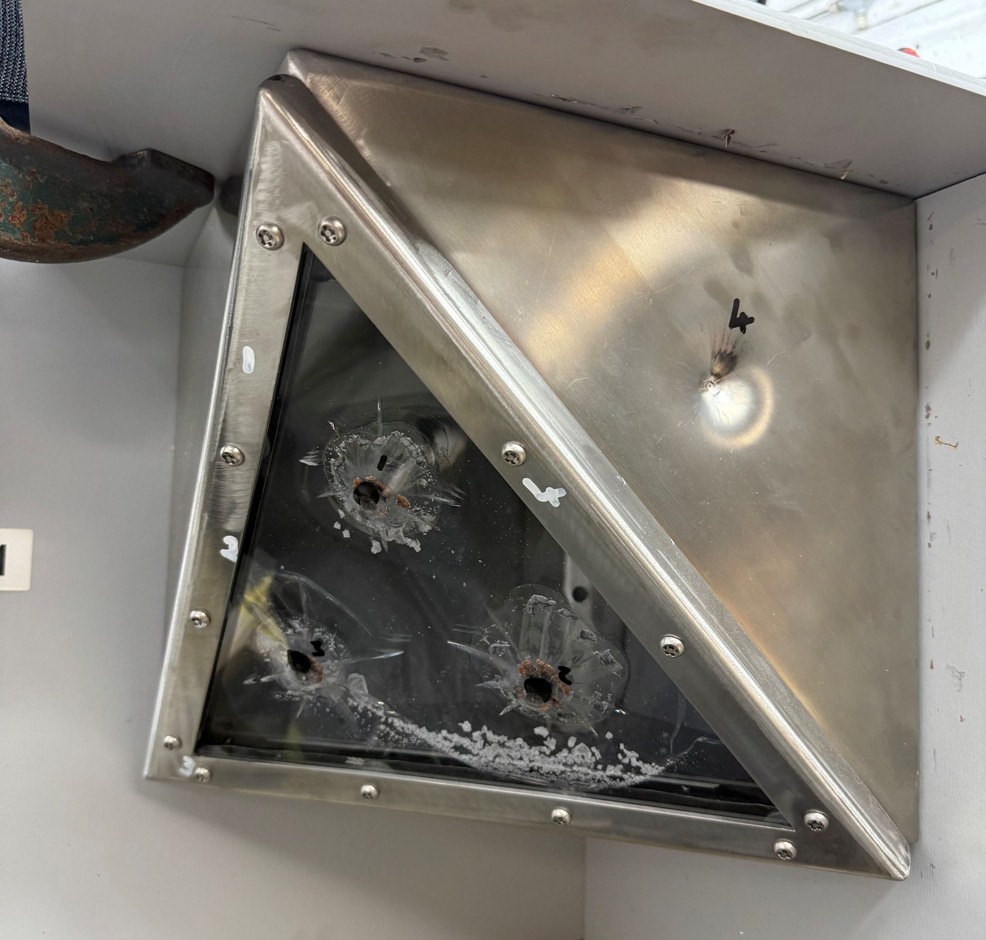 A metal, triangular corner housing features a glass pane with three clear bullet impact holes.