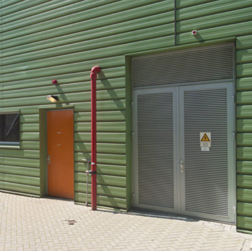 A green metal-sided building featuring an orange door, a red vertical pipe, and double doors with an electrical hazard sign.