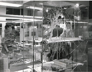 A black-and-white view of a shattered glass display case in a retail setting, with a person visible in the background.